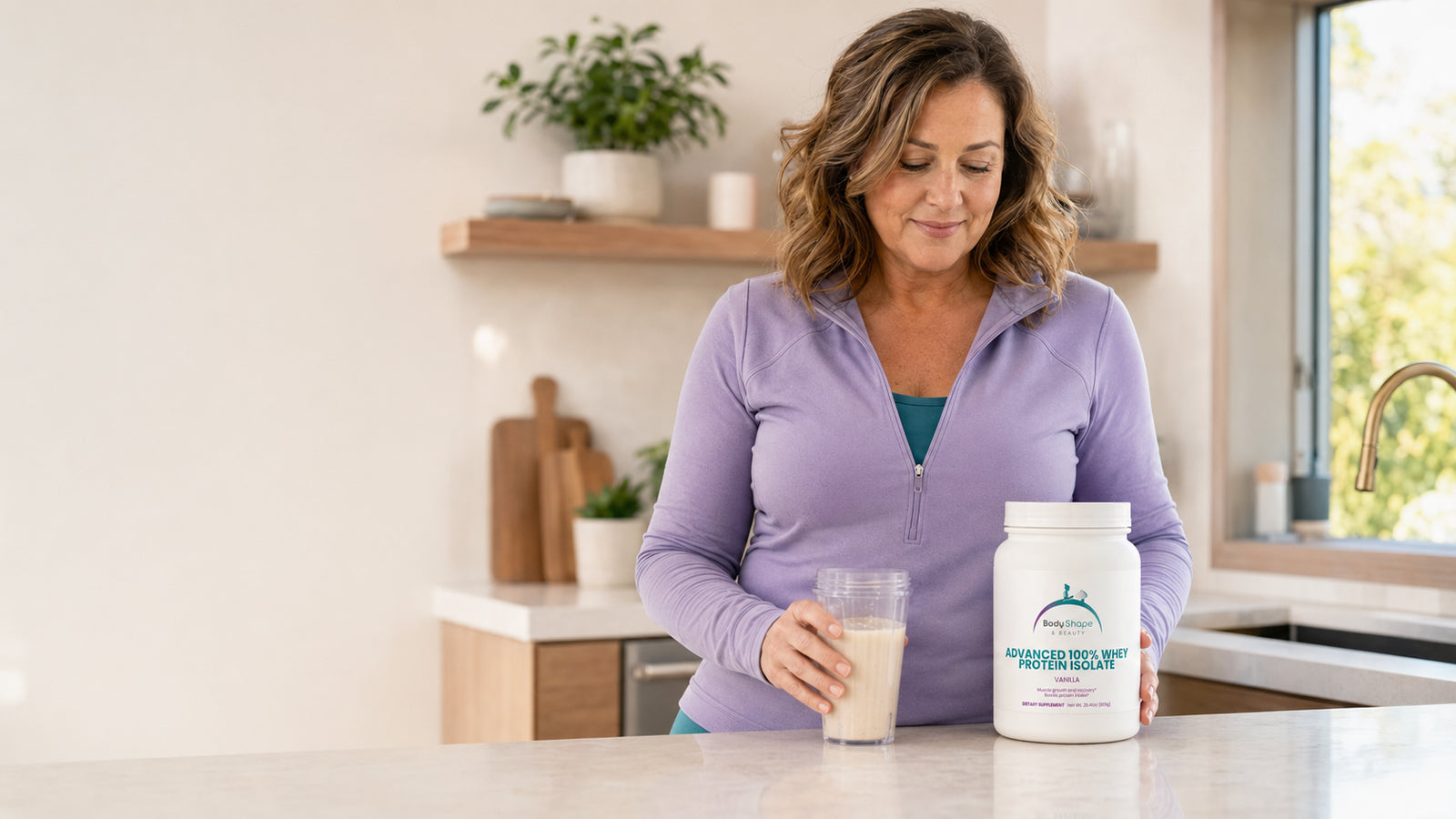 Woman over 40 preparing a vanilla protein shake with Body Shape & Beauty Advanced Whey Protein Isolate in a bright modern kitchen.